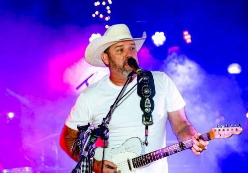 A man wearing a cowboy hat and white T-shirt plays an electric guitar and sings into a microphone on a colorful, smoke-filled stage.