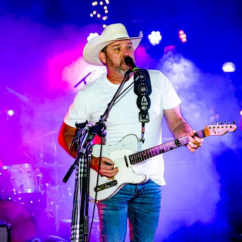 A man wearing a cowboy hat and white T-shirt plays an electric guitar and sings into a microphone on a colorful, smoke-filled stage.