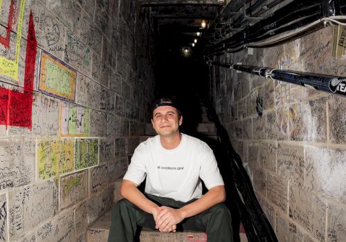 A person sits on steps in a graffiti-covered, narrow passageway with utility cables running along one side and lights overhead, smiling at the camera.