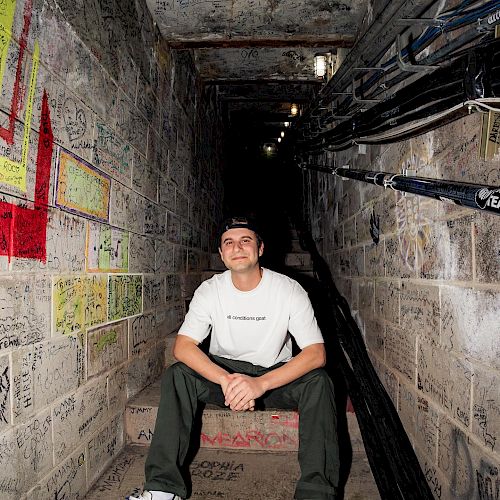 A person sits on steps in a graffiti-covered, narrow passageway with utility cables running along one side and lights overhead, smiling at the camera.