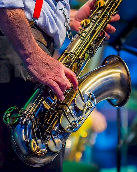 A person holding a saxophone, dressed in a shirt with red suspenders, performing on stage with a blurred background.