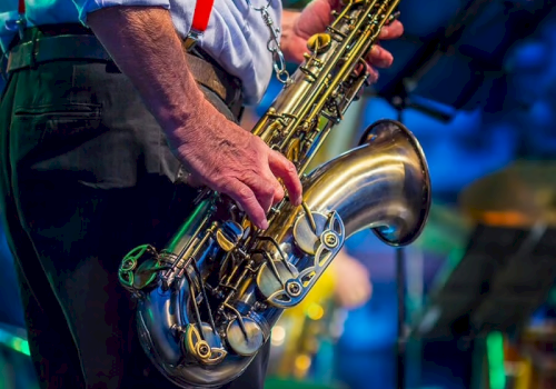 A person holding a saxophone, dressed in a shirt with red suspenders, performing on stage with a blurred background.