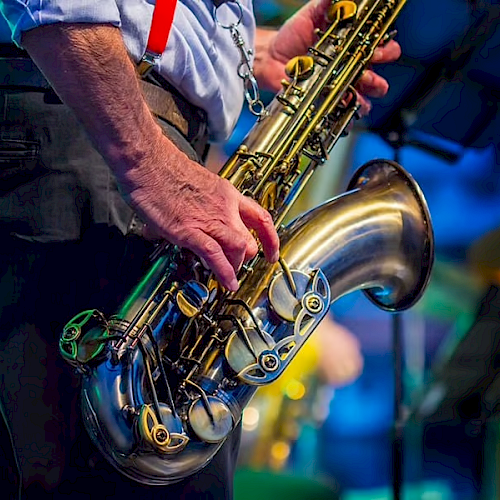 A person holding a saxophone, dressed in a shirt with red suspenders, performing on stage with a blurred background.