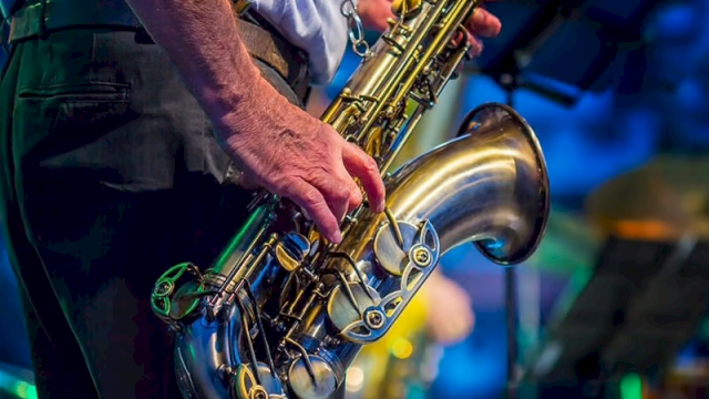 A person holding a saxophone, dressed in a shirt with red suspenders, performing on stage with a blurred background.