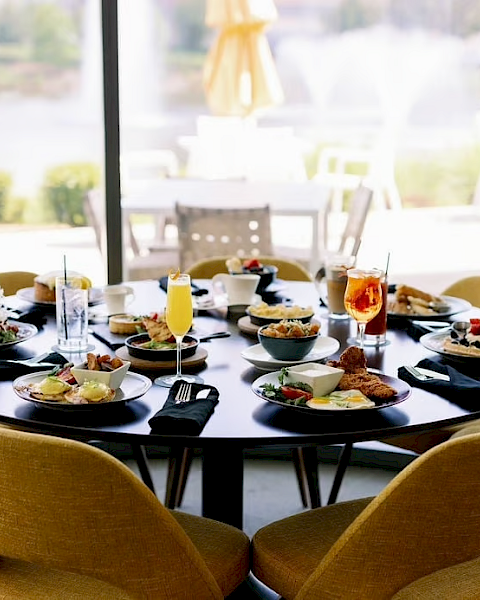 A round dining table set for six with plates of food and beverages, located indoors near large windows with an outdoor view.