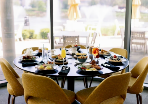 A round dining table set for six with plates of food and beverages, located indoors near large windows with an outdoor view.