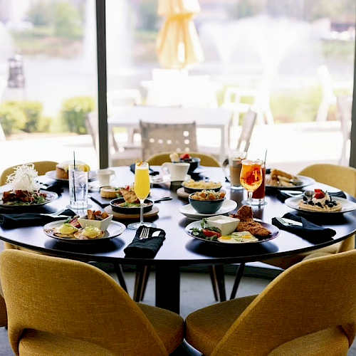 A round dining table set for six with plates of food and beverages, located indoors near large windows with an outdoor view.