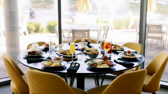 A round dining table set for six with plates of food and beverages, located indoors near large windows with an outdoor view.