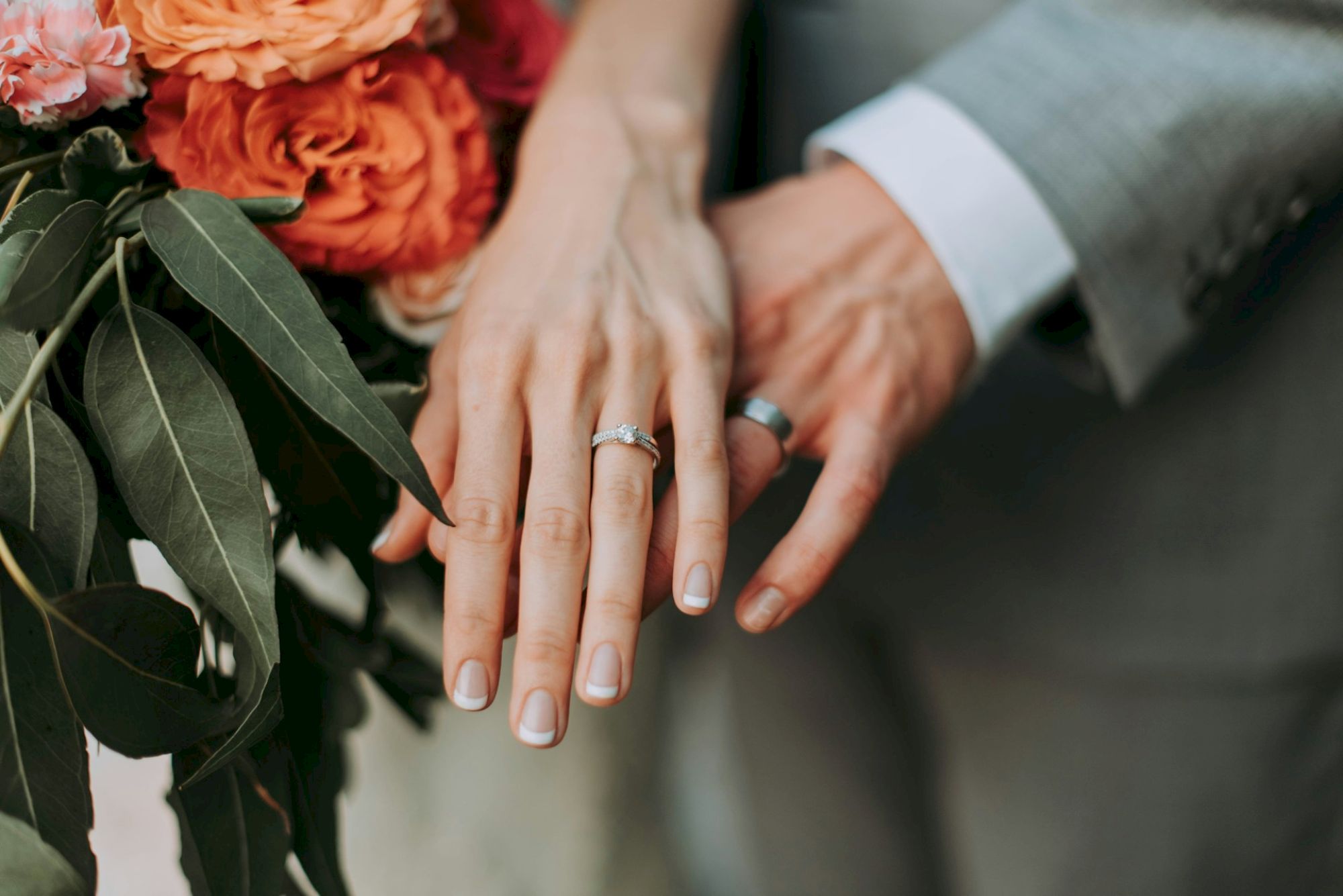 The image shows a close-up of two people wearing wedding rings, with one person's hand on top of the other, next to a bouquet of orange roses and green leaves.