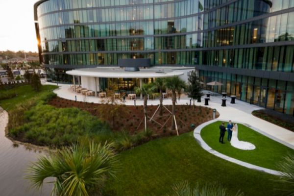 A modern building with a curved glass facade stands by a landscaped garden and a small pond; a couple poses for photos on a grassy lawn.