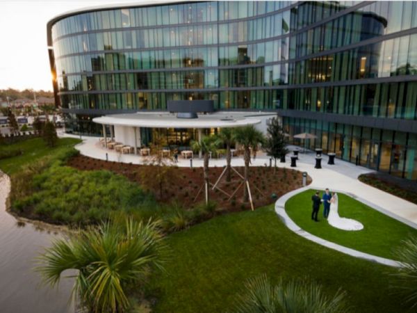 A modern building with a curved glass facade stands by a landscaped garden and a small pond; a couple poses for photos on a grassy lawn.