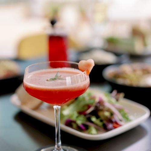 A fancy glass of red wine with a small drink stirrer, served beside a plate of grilled meat and vegetables on a table.