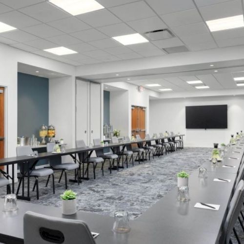 A conference room setup with U-shaped tables, chairs, notepads, water glasses, small plants, a large screen on the wall, and doors in the back.