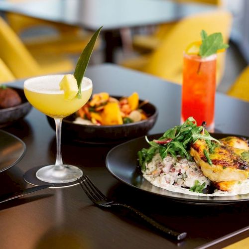 A table set with two cocktails and plates of food, including a main dish of grilled chicken with rice, a side of roasted vegetables, and bread.