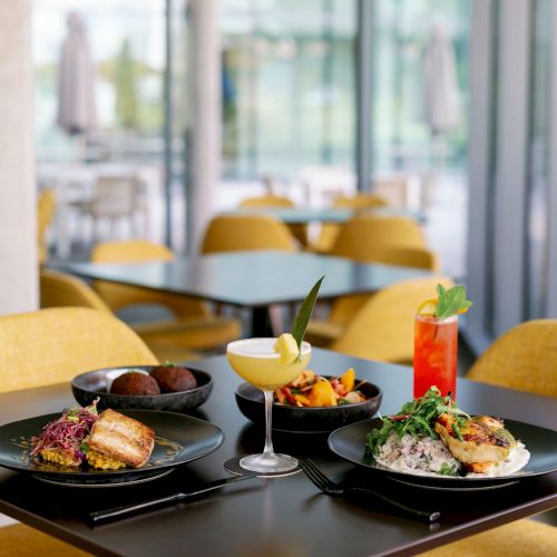 A cozy dining setup with a two-person table, yellow chairs, candles, red napkins, and plated meals by a sunny window.