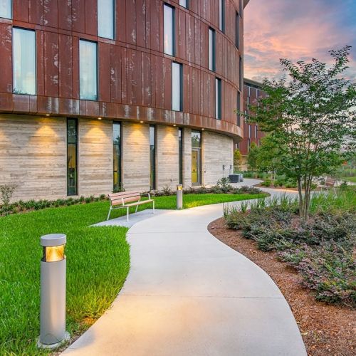 A modern building with a curved design stands beside a landscaped path, featuring grass, plants, a bench, and lit pathway lights at sunset.