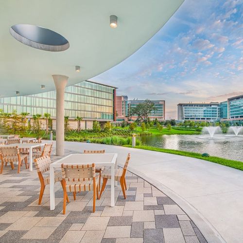 An outdoor patio with tables and chairs overlooks a serene pond with fountains, surrounded by modern buildings under a partly cloudy sky.