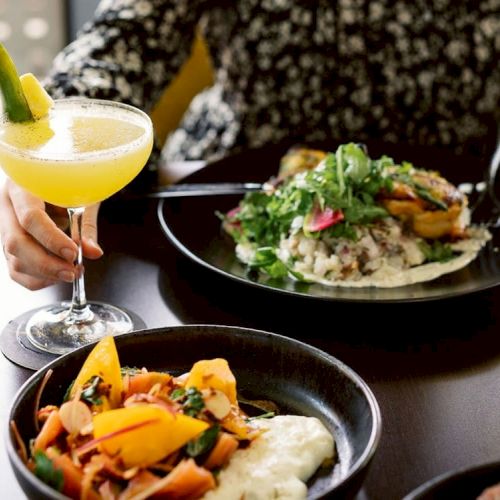 A person with a patterned shirt enjoys a meal featuring a mango drink, a plate of rice and greens, and a bowl of vegetables with sauce.