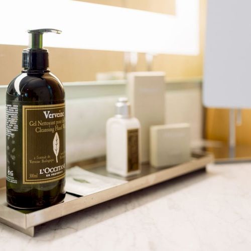 The image shows a bathroom countertop with various toiletries, including a bottle of L'Occitane Verveine Cleansing Hand Gel, soap, and a mirror.