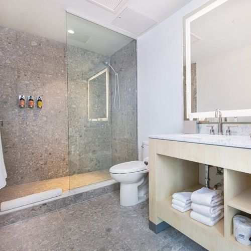 This is a modern bathroom with a glass-enclosed shower, a toilet, a vanity with a large mirror, towels, and toiletries on shelves.