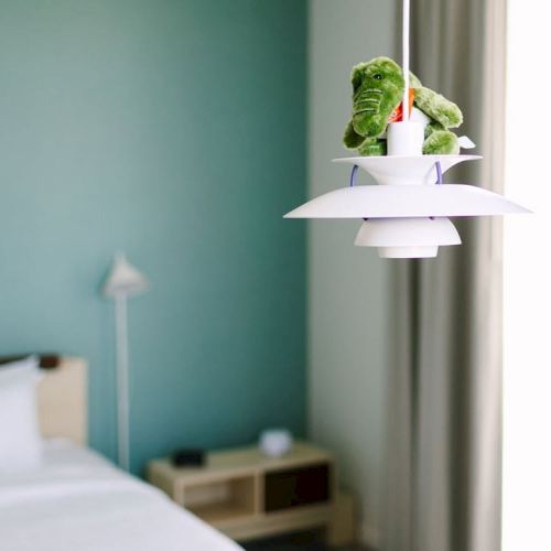 A bedroom with a green wall, white bed, bedside table, and a hanging light fixture with a green stuffed toy on top of it.