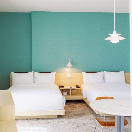 A modern bedroom features two white beds against a teal accent wall, a small round table with chairs, a wooden nightstand, and a pendant light.