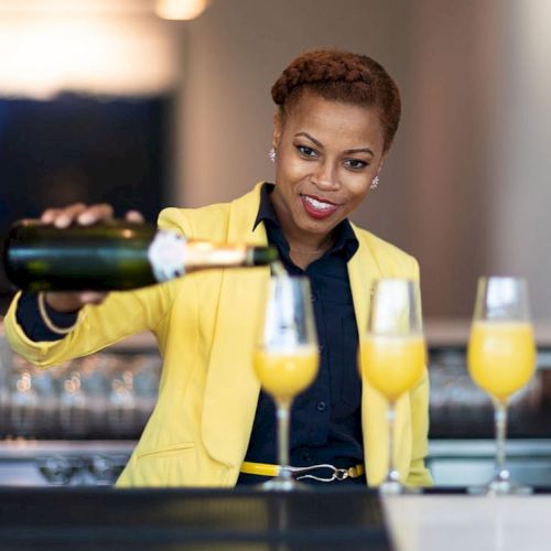 A person wearing a yellow jacket is pouring a beverage into three glasses at a bar or restaurant setting.