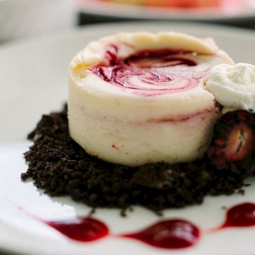 The image shows a small dessert with a swirled topping, sitting on a bed of chocolate crumbs with a dollop of cream and berries.