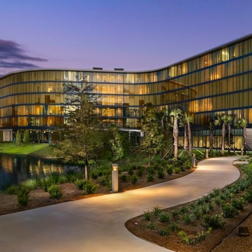 A modern, multi-story building with large glass windows is situated near a water body with a landscaped path leading to it, during twilight.