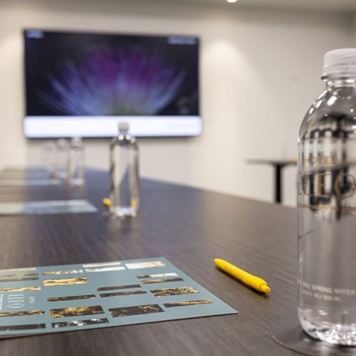 The image shows a conference room set up with bottled water, a yellow pen, and a brochure on a table, with a screen displaying an image in the background.