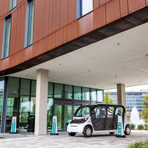 An electric vehicle is parked under a modern building's awning near charging stations. The structure features large glass windows and columns.