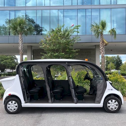 The image shows a white, elongated electric vehicle with its side doors open, parked in front of a modern building with large windows.