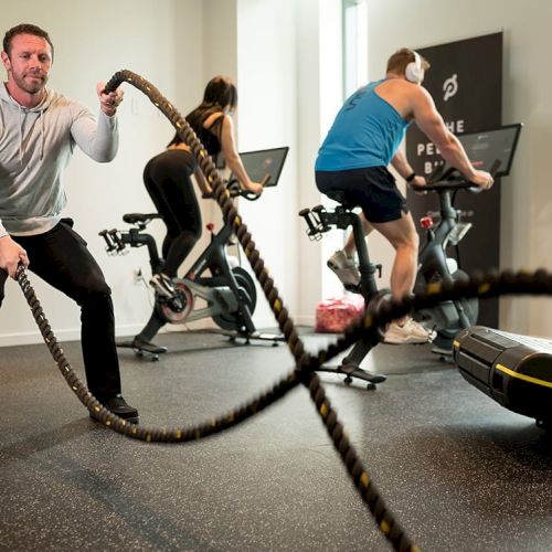 Three people are working out in a gym, one using battle ropes, while two others are riding stationary bikes, and a treadmill is visible.