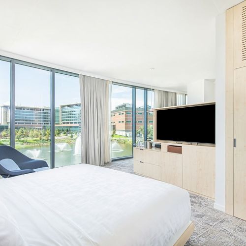 A modern, bright bedroom with a large bed, TV, a blue chair, and floor-to-ceiling windows showcasing a view of buildings and greenery outside.