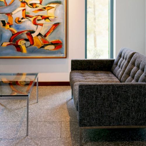 A modern interior with a gray sofa, a glass coffee table, and an abstract painting on the wall. There is a window providing natural light.