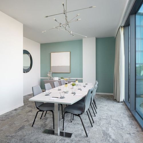 The image shows a modern conference room with a white table, gray chairs, a large window, and minimalistic decor including a wall mirror and artwork.