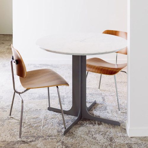 The image shows a small round table with a white tabletop and a metal pedestal base, paired with two wooden chairs. The setting appears minimalist.