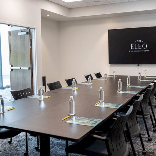 A conference room with a long table, chairs, water bottles, notepads, and a display screen reading 