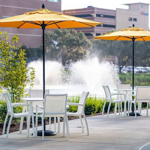 An outdoor cafe area with white chairs and tables, yellow umbrellas, a water fountain in the background, and buildings in the distance.