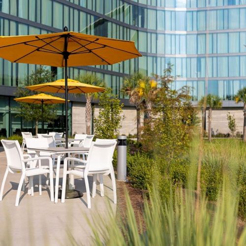 The image shows an outdoor seating area with white chairs and tables under yellow umbrellas, adjacent to a modern glass building and landscaped greenery.