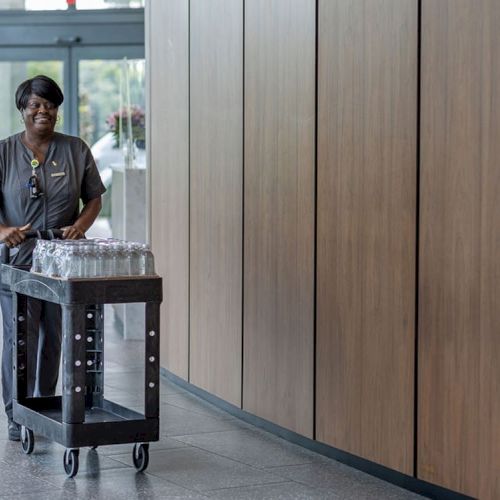 A person in uniform pushes a cart with several glasses in a hallway. The walls are wooden, and the floor is made of tiles.