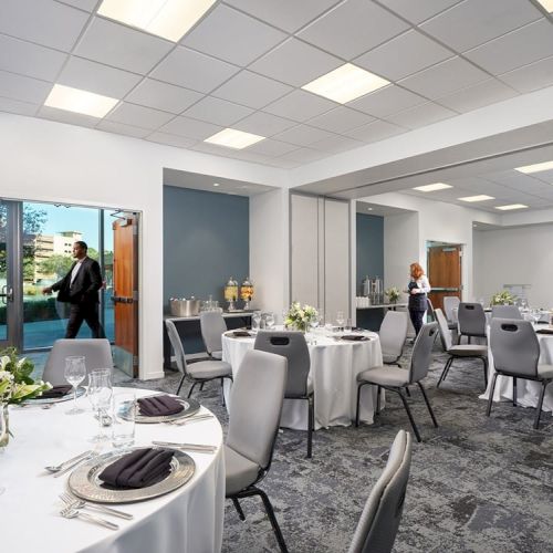 A banquet-style room with round tables set for a formal event, featuring white tablecloths, floral centerpieces, and a few people preparing the setup.