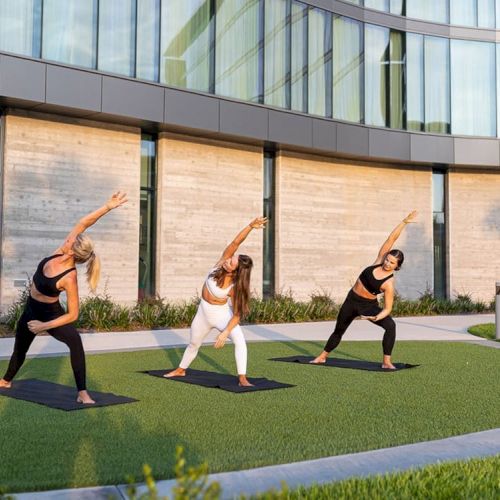 Three individuals are performing yoga on mats outdoors in front of a modern building with curved glass windows and concrete walls.