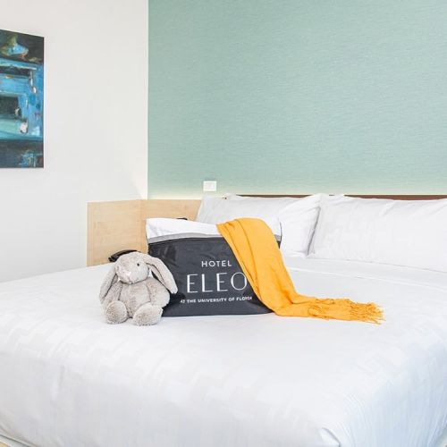 A neatly made bed with white linens, a yellow scarf, a gray stuffed toy, and a sign reading 