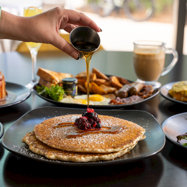 A table of brunch plates: pancakes with berry topping in the center, syrup being poured over them, plus salads, fries, toast, and drinks on the side&mdash;everything ready for a feast.
