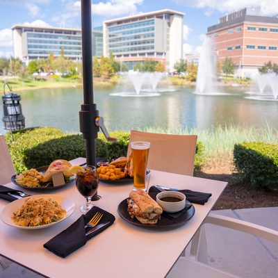 A sunny outdoor dining scene by a pond with fountains, featuring plates of pasta, bread, desserts, beers, and coffee on a white table.
