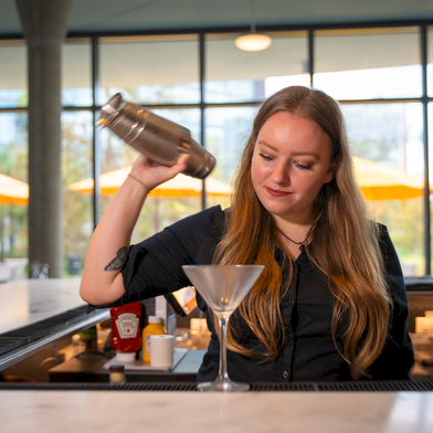 A bartender pours a drink into a martini glass at a modern bar, with a shaker in hand and warm outdoor seating visible outside.