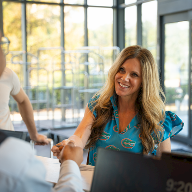A woman smiles and shakes hands with a man across a desk in a bright office lobby.