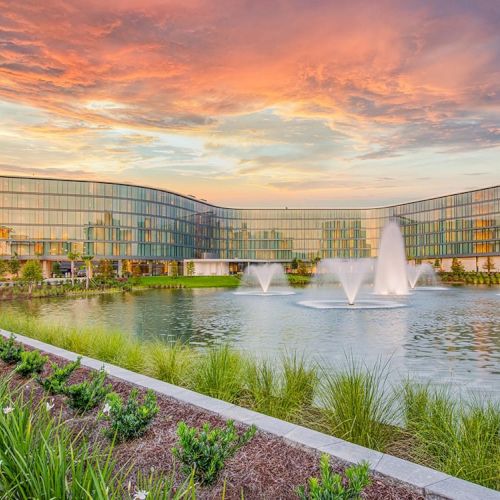 The image shows a modern building with large glass windows, fountains in front of it, and a sunset sky in the background, ending the sentence.