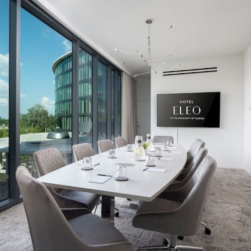 A modern conference room features a long table with chairs, setup for a meeting. A TV on the wall displays 